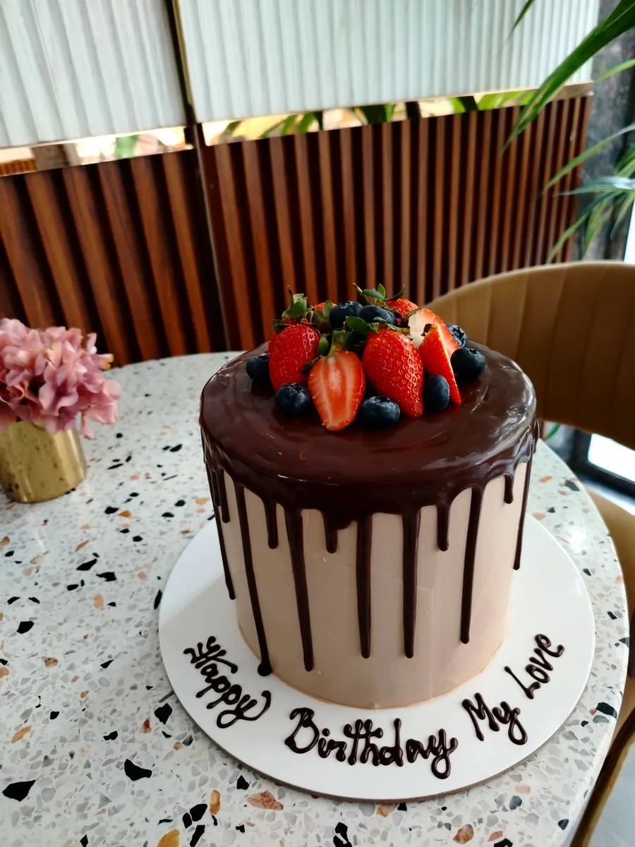 A tall cake with chocolate drip, strawberries and blueberries, Happy Birthday My Love piped on the board